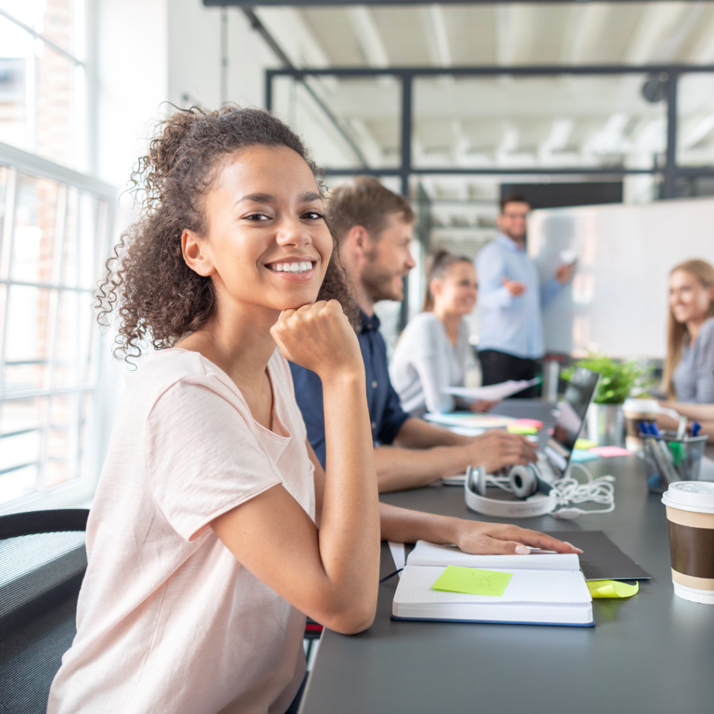 A young black female apprentice is sitting next to a colleague in front of a computer. She is smiling at the camera whilst doing online learning for her Level 3 Team Leader Supervisor apprenticeship