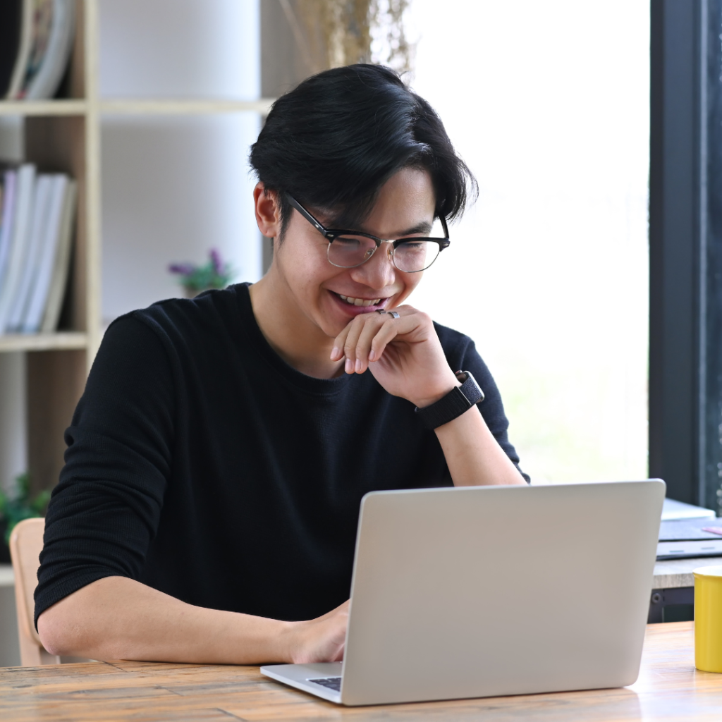 A young asian male apprentice wearing glasses is sitting in front of a silver laptop. He is smiling whilst working on a project for his Level 3 Team Leader Supervisor apprenticeship